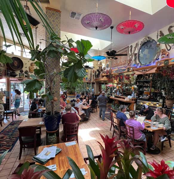 Bustling restaurant interior with eclectic global decor, hanging parasols, lush plants, and guests enjoying meals in a vibrant space.