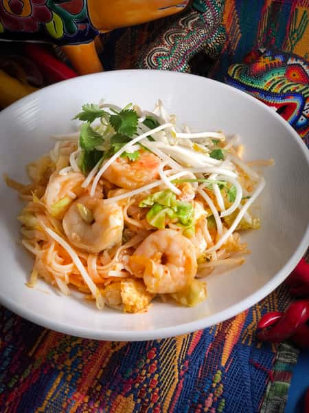 Shrimp pad Thai with rice noodles, bean sprouts, cabbage, egg, and cilantro, served in a white bowl on a colorful textile.