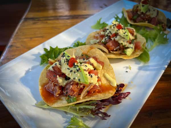 Ahi poke tostadas topped with avocado, creamy drizzle, sesame seeds, and greens on a crispy shell, served on a white platter.