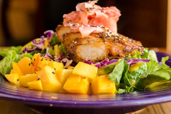 Seared white fish over mixed greens with mango, avocado, cabbage, and pickled ginger, garnished with sesame seeds.