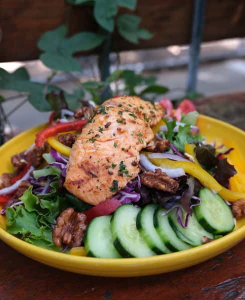 Grilled salmon salad with mixed greens, cucumbers, bell peppers, candied walnuts, and red onions, served in a vibrant yellow bowl.