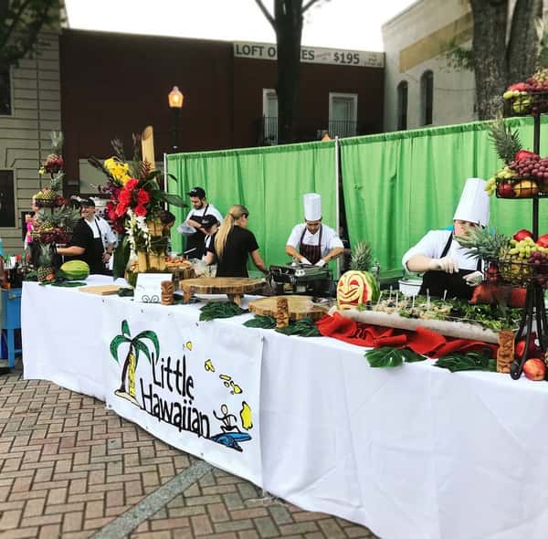 Outdoor food booth for "Little Hawaiian" with chefs preparing dishes, tropical fruits, and carved watermelon.