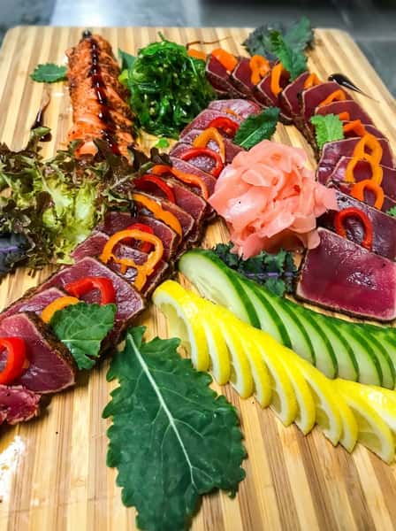 Seared tuna sashimi with sweet peppers, salmon, seaweed salad, pickled ginger, lemons, and cucumbers on a wooden board.