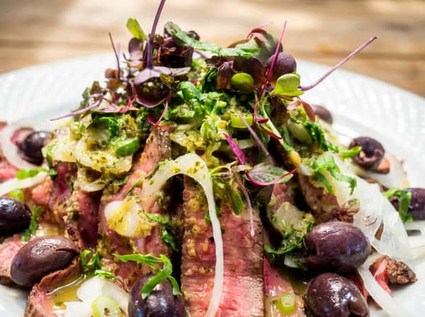 Sliced beef with green sauce, white onions, olives, and microgreens on a decorative plate.