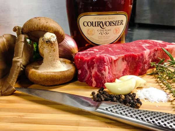 Ingredients for cooking, including a raw steak, mushrooms, shallots, garlic, herbs, peppercorns, and a bottle of Courvoisier cognac.