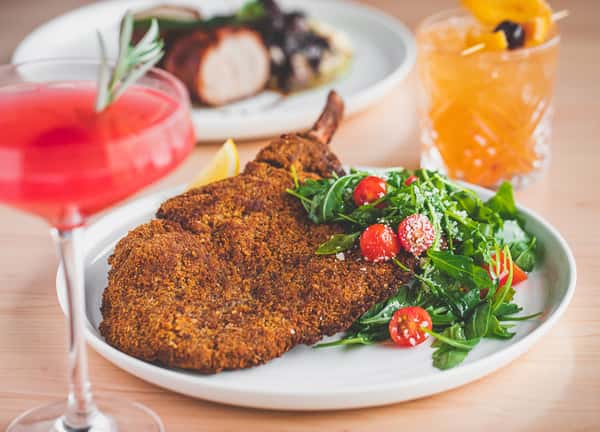 A crispy chicken Milanese topped with fresh arugula and cherry tomatoes, paired with a colorful cocktail, representing the diverse Italian cuisine