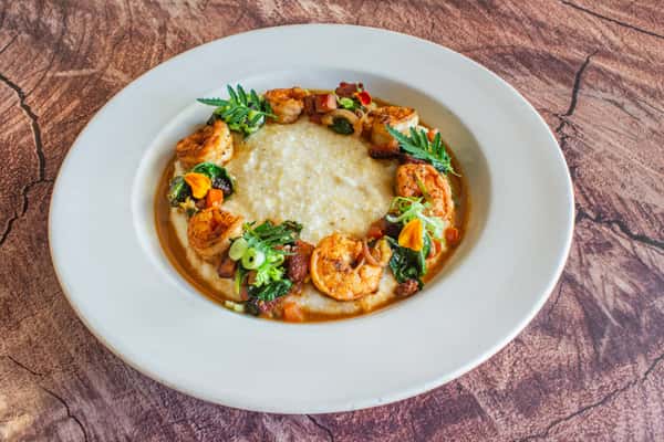 A Southern-style shrimp and grits dish featuring tender sautéed shrimp in a rich, savory sauce served over creamy stone-ground grits. Garnished with fresh herbs, this comforting entrée delivers bold Cajun-inspired flavors and a beautifully refined presentation.