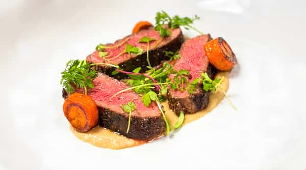 Three slices of medium-rare steak on a bed of puree, garnished with microgreens and roasted carrots.