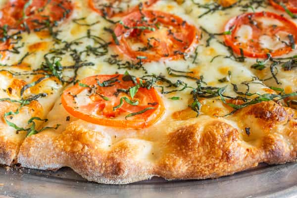 A close-up of a handcrafted Mediterranean-style Margherita pizza topped with melted mozzarella, fresh tomato slices, aromatic herbs, and a golden, wood-fired crust. The crisp edges and vibrant toppings highlight the pizza's fresh ingredients and authentic Italian flavor.