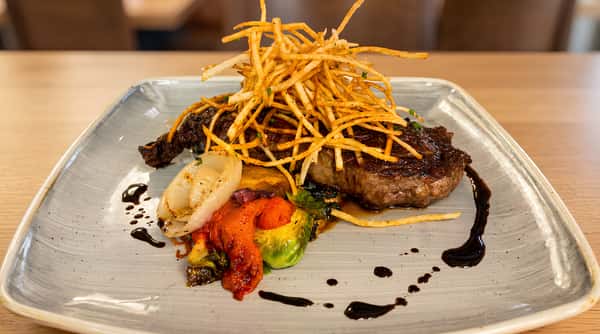 Grilled steak, topped with crispy shoestring fries, served with roasted vegetables (onions, red peppers, Brussels sprouts), and drizzled with a dark sauce on a gray plate.