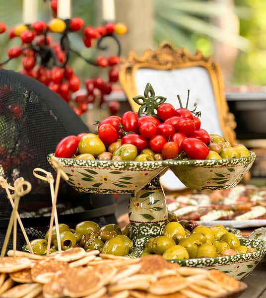 Buffet table with a tiered bowl of cherry tomatoes and olives, a dish of small pancakes, and a person's hand reaching for food.