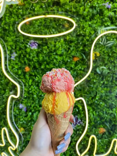 Hand holding waffle cone with pink and mango ice cream, sprinkles and syrup, against neon angel wings wall