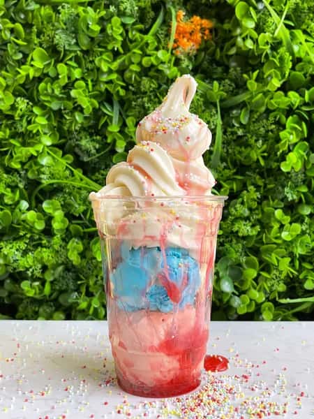 Colorful sundae with blue and pink ice cream, whipped cream, sprinkles and red syrup in clear cup on green wall backdrop