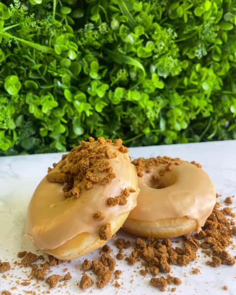 Two glazed donuts topped with biscuit crumbs on white surface against lush green leafy backdrop