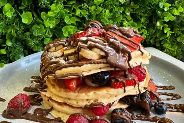 Stack of fluffy pancakes layered with strawberries and blueberries, drizzled in chocolate sauce and dusted with powdered sugar