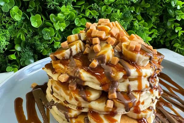 Stack of pancakes topped with banana slices, caramel cubes, chocolate and caramel drizzle on a gray plate against green foliage