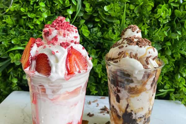 Two loaded milkshakes topped with soft serve—one strawberry with fresh slices, one caramel chocolate with cookie crumbles.