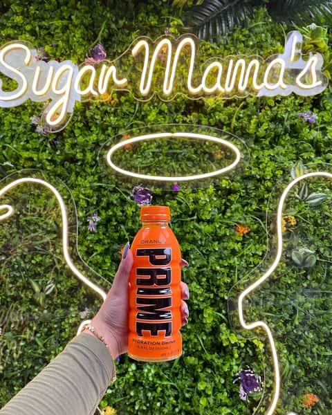 This image shows a hand holding an orange bottle of PRIME Hydration Drink (Orange flavor) in front of a vibrant green plant wall with a neon "Sugar Mama's" sign and glowing halo-style outlines in the background.