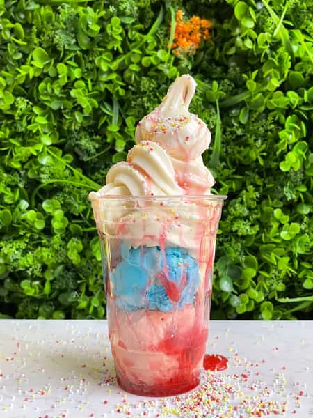 Colorful ice cream sundae with blue and pink scoops, vanilla swirl, sprinkles, and red syrup in clear cup against green wall