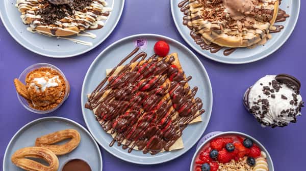 Assorted dessert plates with chocolate-drizzled crepes, waffles, churros, ice cream, and fresh berries on purple table