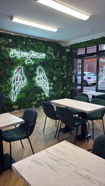 Modern café interior with marble tables, green chairs, and neon angel wings on a lush plant wall reading Sugar Mama's