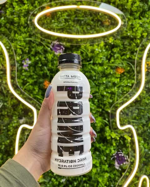 Hand holding PRIME Meta Moon hydration drink in front of neon angel wings on green foliage wall