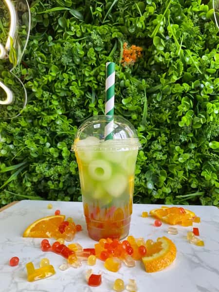 Clear cup of green iced drink with striped straw, citrus slices, and jelly candies on table before leafy backdrop