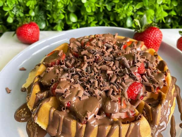 Waffle topped with strawberries and thick chocolate drizzle, covered in chocolate curls on a gray plate with greenery backdrop.