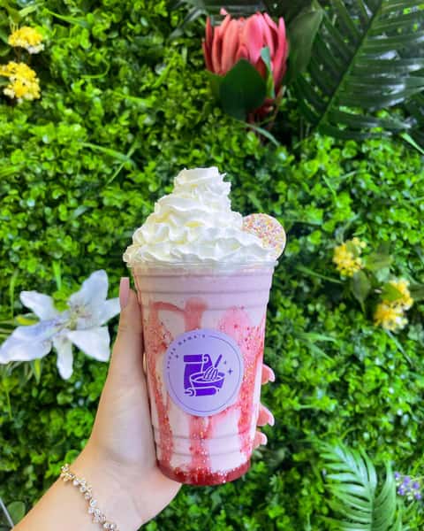 Hand holding pink milkshake with whipped cream and sprinkles in Sugar Mama's cup against lush green wall backdrop