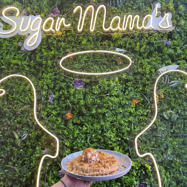 Plate of caramel-drizzled waffle topped with biscuit crumbs held before Sugar Mama's neon sign and green wall