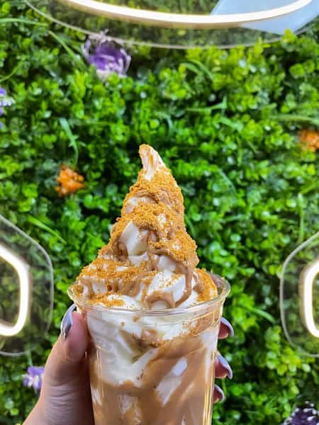 Soft serve sundae drizzled with caramel and cookie crumbs, held up against a lush green wall with neon outline lights.