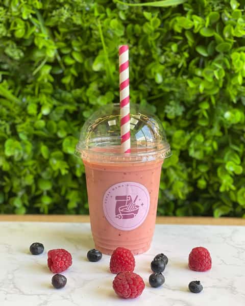 Pink berry smoothie in clear cup with striped straw, raspberries and blueberries on marble surface, green leaf wall backdrop
