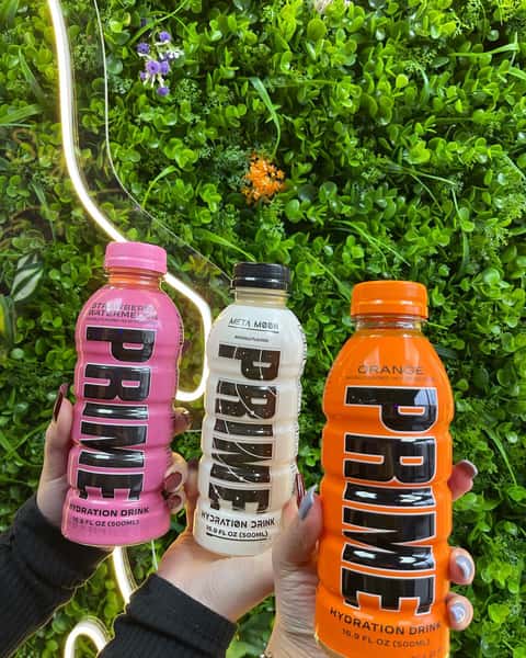 Three hands holding PRIME hydration drinks in strawberry watermelon, Meta Moon, and orange flavors against green wall