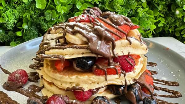 Stack of fluffy pancakes layered with strawberries and blueberries, drizzled in chocolate sauce and dusted with powdered sugar