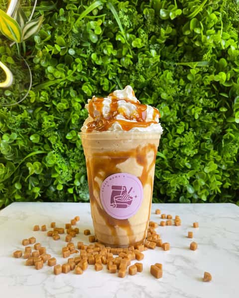 Caramel milkshake topped with whipped cream and caramel drizzle, surrounded by caramel cubes on white surface