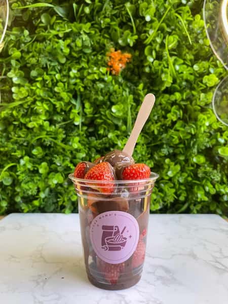 Clear cup of chocolate-covered strawberries with wooden spoon, set against lush green wall backdrop