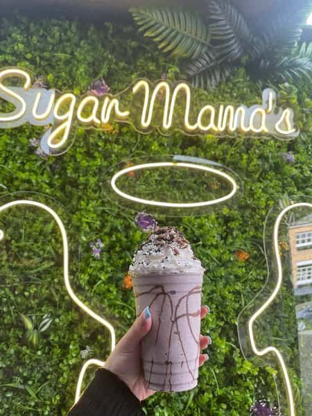 Hand holding chocolate drizzle milkshake with whipped cream before Sugar Mama's neon sign and leafy green wall