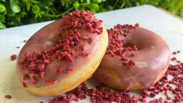 Two glazed donuts topped with crushed freeze-dried strawberries on white surface, green foliage background