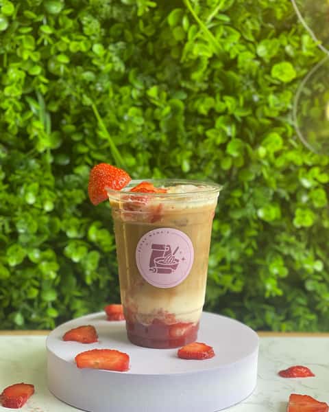 Iced strawberry latte in clear cup with fresh strawberry slices on white stand, green leafy wall backdrop