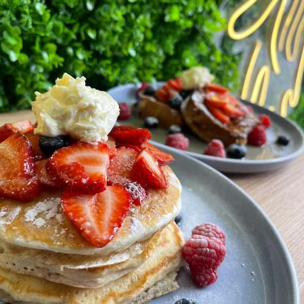 Stack of fluffy pancakes topped with fresh strawberries and cream, raspberries on plate, café setting background