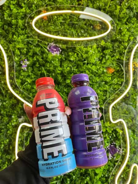 Hand holding red, white and blue PRIME Ice Pop and purple PRIME Grape hydration drinks before neon angel wings and green wall backdrop