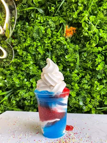 Red, white and blue layered drink topped with vanilla soft serve, sprinkles on table, green wall backdrop