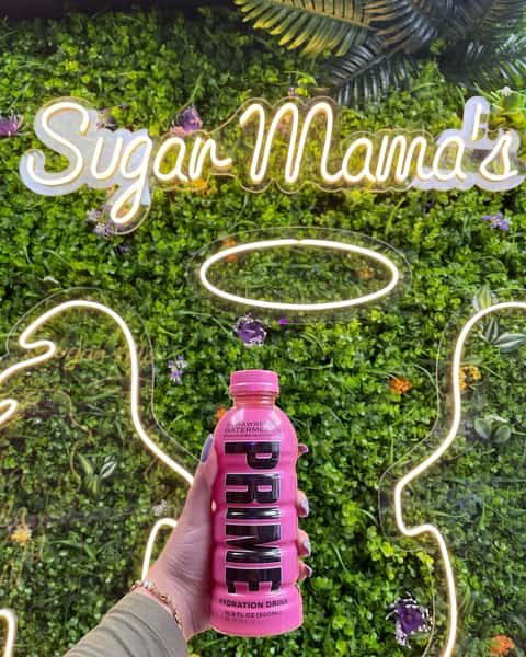 Hand holding pink PRIME Strawberry Watermelon hydration drink before Sugar Mama's neon sign and green wall backdrop