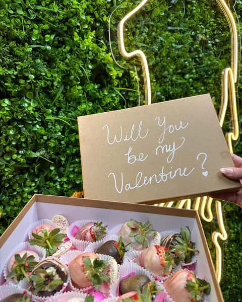 Open gift box of chocolate-covered strawberries with note reading "Will you be my Valentine?" against green wall backdrop