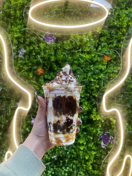Hand holding caramel and chocolate sundae topped with whipped cream and chips before neon angel wings on green wall