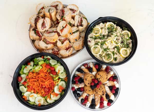 Top-down view of an Italian catering spread with mini hoagie sandwiches, lemon herb couscous, crisp garden salad, and a tray of desserts including cannoli and cookies.