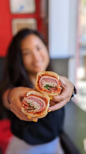 Smiling woman presenting an Italian hoagie sliced in half, showcasing layers of cured meats, cheese, lettuce, and tomato on fresh bread, inside a deli.