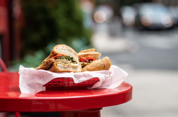 Close-up of a toasted Italian sandwich filled with fresh ingredients, served in a red basket on a red café table outside an urban sandwich shop.