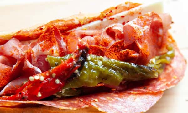 Bold close-up of a spicy Italian sandwich loaded with capicola, salami, roasted red and green peppers, and sprinkled with red chili flakes.