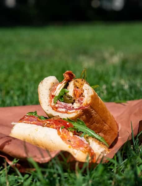 Hearty Italian hoagie sandwich featuring cured meats, provolone, arugula, and hot peppers on a hoagie roll, styled for an outdoor picnic setting.